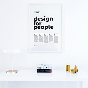 May include: A white desk with a white lamp, a framed poster that says "design for people", and two books stacked on top of each other. The top book is titled "The Book of Symbols". There are three gold metal decorative objects on the desk.