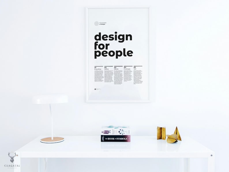 May include: A white desk with a white lamp, a framed poster that says "design for people", and two books stacked on top of each other. The top book is titled "The Book of Symbols". There are three gold metal decorative objects on the desk.