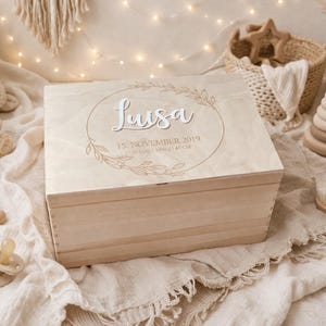 Könnte beinhalten: Eine hellfarbene Holzkiste mit dem Namen "Luisa" in Schreibschrift, umgeben von einem Kranzdesign. Die Kiste ist von Babyspielzeug umgeben, darunter ein Teddybär, ein regenbogenförmiges Spielzeug und ein Schnuller. Der Text "15. November 2019" ist ebenfalls sichtbar.