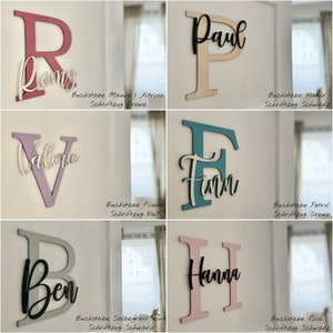 May include: A collection of wooden letter signs in various colors, including mauve, natural, teal, and pink. Each sign features a large letter with a smaller name overlay. The signs are mounted on a white wall, showcasing a personalized decor theme.