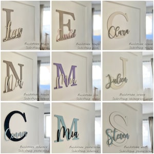 May include: A collection of wooden letter cutouts in various colors, each with a name in a contrasting script font. The letters are mounted on a white wall. The letters are in taupe, lilac, black, and white.