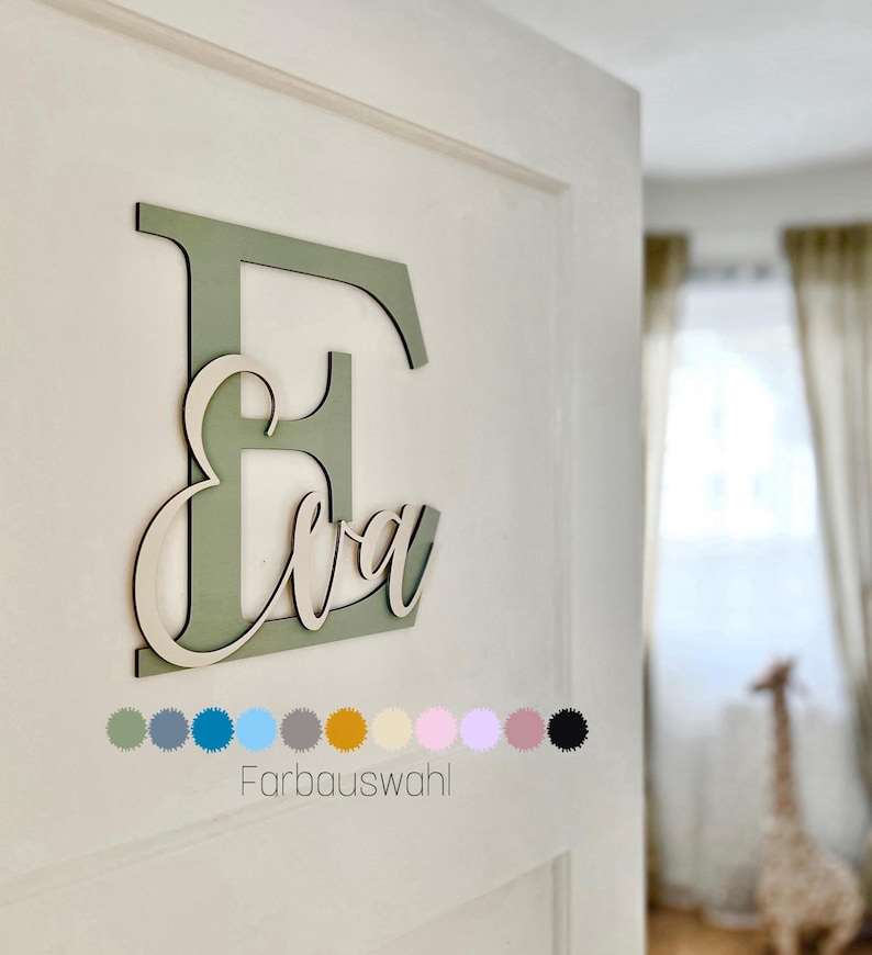 May include: A wooden wall decoration featuring the name "Eva" in a cursive font, layered over a large letter "E". The letter "E" is painted in a muted green, with the name in a natural wood color. The image includes a color palette selection.