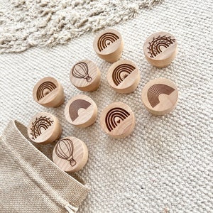 Op de afbeelding: Een set van 10 houten stempels met verschillende ontwerpen, waaronder regenbogen, luchtballonnen en zonsopgangen. De stempels zijn gerangschikt op een lichtbeige oppervlak met een kleine jute zak in de linkerbenedenhoek.