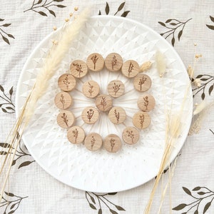 Op de afbeelding: Een set van 16 houten bloemstempels, gerangschikt in een cirkel op een wit bord. De stempels hebben allemaal verschillende ontwerpen en bevatten verschillende bloemen en bladeren.