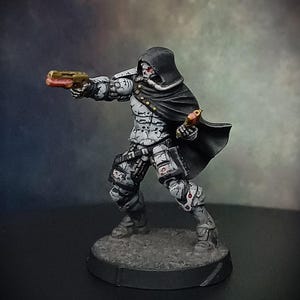 May include: A white and gray painted miniature figure of a person wearing a black hooded cloak and holding a gun in one hand and a small bird in the other. The figure is standing on a round black base.