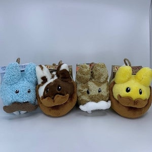 May include: Four plush bunny toys in various colors: light blue, brown and white, tan, and yellow. Each bunny has a brown mustache and a small loop at the top. The bunnies are sitting on top of small cardboard boxes.