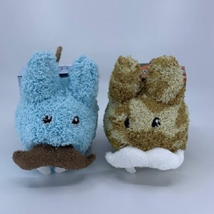 May include: Two plush toys shaped like bunnies with mustaches. One bunny is light blue with a brown mustache and the other is brown with a white mustache.