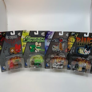 May include: Five DC Comics character action figures in packaging. The figures are Green Lantern, Cyborg, Raven, Red Hood, and a figure with a red and black costume. Each figure is in a plastic blister pack with a colorful background and the DC Comics logo.