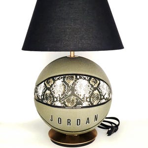 以下が含まれることがあります： バスケットボールのデザインと黒いランプシェードが特徴的なユニークなランプ。バスケットボールにはヘビ革模様と「JORDAN」の文字がプリントされています。ランプはオリーブグリーンで、金色のベースが付いています。