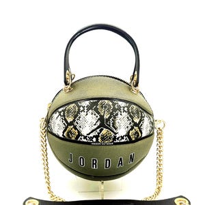Könnte beinhalten: Olivgrüne Handtasche in Basketballform mit schwarzem Griff und goldener Kette. Die Tasche hat ein Schlangenmuster und das Wort "JORDAN" in weißen Blockbuchstaben. Es gibt einen schwarzen Schulterriemen aus Leder.