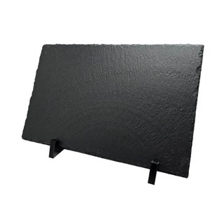 Op de afbeelding: Een zwarte leisteen serveerschaal met twee zwarte metalen standaards. De schaal is rechthoekig en heeft een ruw, getextureerd oppervlak.