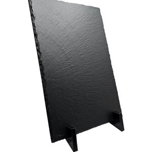 Op de afbeelding: Een zwart leisteen serveerplank met een standaard. De plank is rechthoekig en heeft een ruw, getextureerd oppervlak. De standaard is gemaakt van zwart metaal en heeft twee poten.