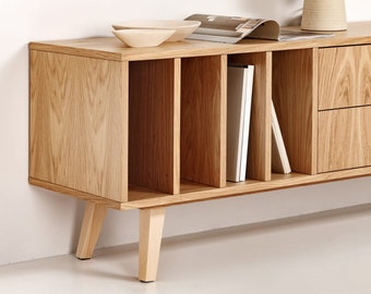 May include: A light wood entertainment center with open shelves and a drawer. The entertainment center has four legs and is sitting on a white surface. A lamp is sitting on a surface behind the entertainment center.