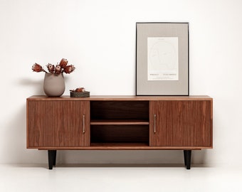 May include: A mid-century modern style wooden cabinet with two doors and a shelf. The cabinet is dark brown with black legs and silver handles. A framed print with a line drawing of two faces is on top of the cabinet. A vase with dried flowers is on the shelf.