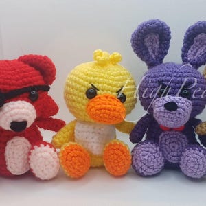 May include: Four handmade crochet plush toys arranged in a row. From left to right: a red fox with a black eye patch, a yellow duck with an orange beak, a purple rabbit with long ears, and a tan bear wearing a black hat and bow tie.