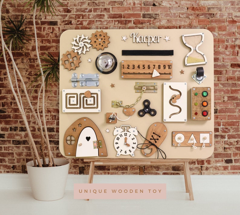 May include: A beige wooden activity board with various textures and activities for children. It features a clock, a door with a latch, a zipper, a gear, a knob, a chain, a spinner, a maze, a number line, a light switch, a shape sorter, and an hourglass. The board is labeled "Kacper" at the top.