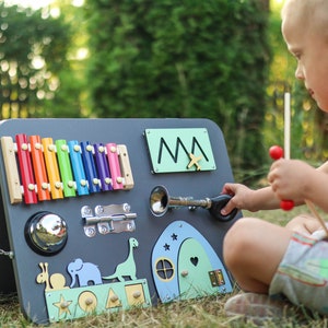 Può includere: Un bambino gioca con un pannello di attività in legno con uno xilofono, una campana, un chiavistello, una porta, un dinosauro, una casa e un classificatore di forme. Il pannello è dipinto di grigio e ha uno schema di colori verde e blu.