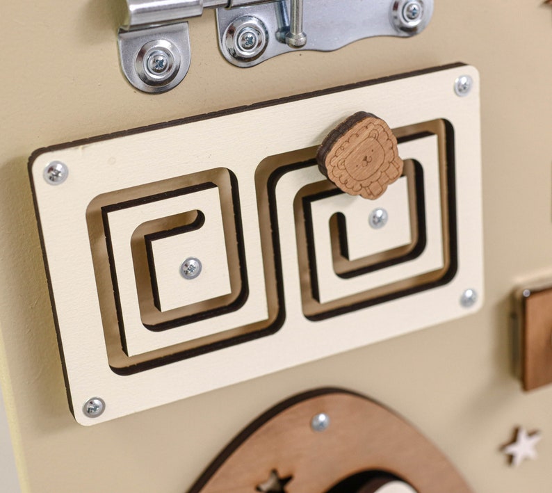 May include: A beige wooden activity board with a variety of textures and shapes for children to explore. The board features a small wooden lion with a brown mane, a latch, and a maze with a sliding piece.