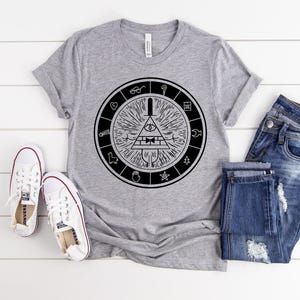 May include: A gray t-shirt with a black graphic of a circular design with an eye, triangle, and other symbols. The shirt is paired with blue jeans and white sneakers. The items are arranged on a white wooden surface.