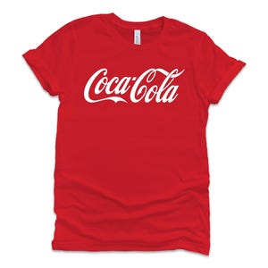 May include: A red t-shirt with the Coca-Cola logo in white script across the front. The shirt has short sleeves and a crew neck. The sleeves are cuffed.