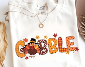 May include: Cream-colored t-shirt with the word "GOBBLE" in orange, brown, and red lettering, featuring a turkey wearing a pilgrim hat. The shirt has a gold necklace and a tag that says "COUNTRY COLORS".