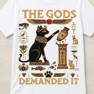 Op de afbeelding: Een wit t-shirt met een grafische print van een zwarte kat versierd met Egyptische sieraden, reikend naar een decoratieve vaas op een voetstuk. De tekst THE GODS DEMANDED IT is in goud gedrukt, omringd door diverse Egyptische symbolen.