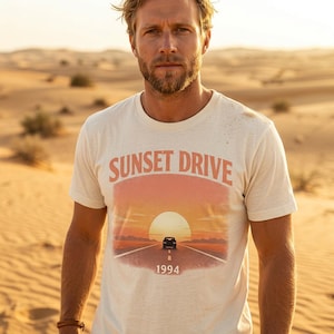 May include: Cream-colored t-shirt with the words "SUNSET DRIVE" above a graphic of a car driving on a road towards a sunset. The year "1994" is printed below the graphic. The shirt is short-sleeved.