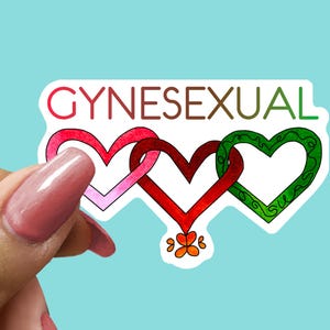 May include: A sticker with the word "GYNESEXUAL" in red, brown, and green. The sticker features three interlocking hearts in pink, red, and green, with a small orange flower design at the bottom. The sticker is held against a light blue background.