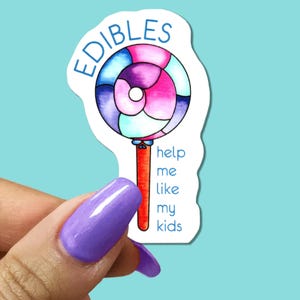 May include: A colorful sticker featuring a lollipop design with the word "EDIBLES" in a blue arc at the top. The lollipop has a red stick and a swirl of pink, purple, and blue. The text "help me like my kids" is written vertically.