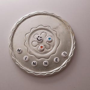 May include: A silver circular coaster with a raised design in the center. The design features a smiley face, two hearts, and a star. The word "MINNES" is spelled out around the edge of the coaster with white letters.