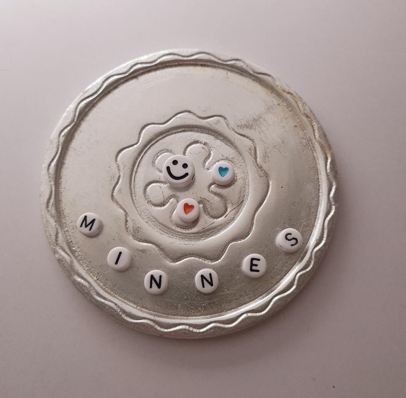 May include: A silver circular coaster with a raised design in the center. The design features a smiley face, two hearts, and a star. The word "MINNES" is spelled out around the edge of the coaster with white letters.