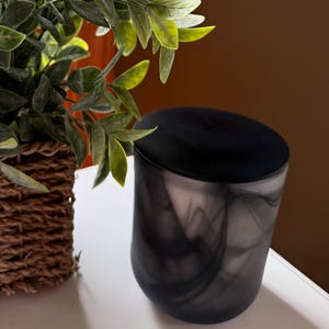 May include: A decorative arrangement featuring a green leafy plant in a woven basket and a black and gray marbled candle holder. The plant has light green leaves and is set against a window and a brown wall.
