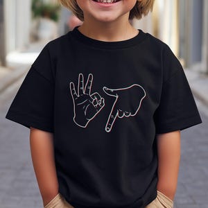 Könnte beinhalten: Schwarzes T-Shirt mit weißem Handgesten-Design. Das Kind trägt khakifarbene Shorts und eine schwarze Sonnenbrille. Das Foto wurde auf einer gepflasterten Straße aufgenommen.
