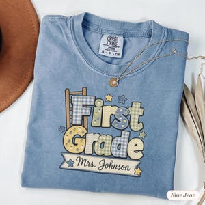 Peut inclure: Un t-shirt bleu à manches courtes avec une impression graphique affichant le texte First Grade et Mrs. Johnson. Le motif comprend une échelle, des étoiles et des lettres aux motifs à carreaux et lignés dans des tons de bleu, jaune et vert.