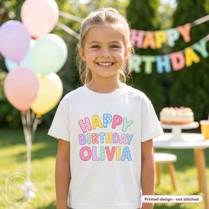 以下が含まれることがあります： 水玉、ストライプ、花柄など様々なパターンでHAPPY BIRTHDAY OLIVIAとカラフルにプリントされた白い半袖Tシャツ。デザインは生地にプリントされており、刺繍ではありません。Tシャツはクルーネックになっています。