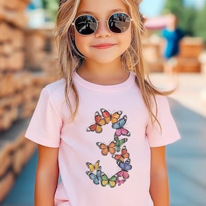 May include: A young girl wearing a pink t-shirt with a butterfly design that forms the number three. She is wearing sunglasses and has long blonde hair.