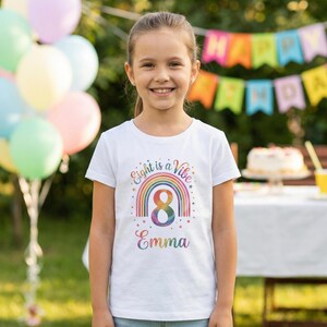Könnte beinhalten: Ein weißes T-Shirt mit einem bunten Regenbogen-Design und dem Text "Eight is a Vibe" und "Emma". Die Zahl 8 befindet sich in der Mitte des Regenbogens. Das Mädchen lächelt vor einer Geburtstagsfeier-Kulisse.