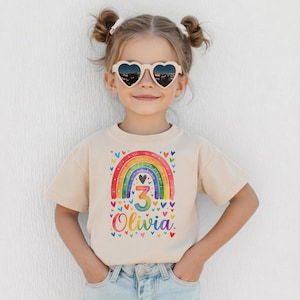 May include: A beige t-shirt with a vibrant rainbow design, the number "3", and the name "Olivia". The rainbow is framed by small hearts. The child is wearing heart-shaped sunglasses.