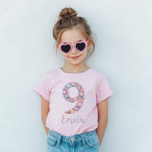 May include: A light pink t-shirt with a seashell-decorated number 9 and the name "Emma" in a cursive font. The girl is wearing pink heart-shaped sunglasses and blue jeans. The girl has her hair up in a bun.