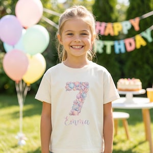 May include: A cream-colored t-shirt with a seashell-themed number 7 design and the name "Emma" in a cursive font. The shirt is worn by a smiling child, with balloons and a birthday banner in the background.