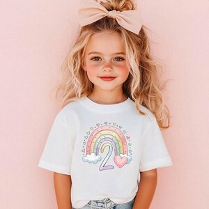 May include: A white t-shirt featuring a colorful rainbow design with the number "2" and a heart. The rainbow includes pastel colors and is decorated with small hearts. The shirt is worn by a young child with blonde hair and a large bow.
