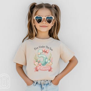 May include: A beige short-sleeved t-shirt featuring a graphic of a teal mermaid-tail number five, a pink bow, seashells, and seaweed. The text reads Five Under The Sea and the name Olivia on a pink banner. The shirt is worn with light blue jeans.