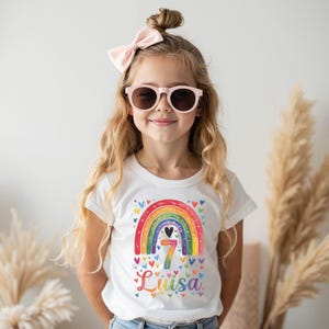 Op de afbeelding: Een wit t-shirt met een kleurrijk regenboogontwerp, het nummer 7 en de naam "Luisa". De regenboog is versierd met hartjes. Het t-shirt wordt gedragen door een jong persoon met een roze zonnebril en een roze strik.