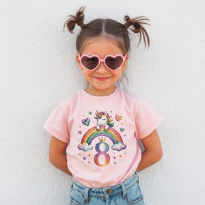 Könnte beinhalten: Ein junger Mensch trägt ein rosa T-Shirt mit einer bunten Grafik eines Einhorns auf einem Regenbogen, der Zahl 8 und Herzen. Die Person trägt auch eine rosa herzförmige Sonnenbrille und blaue Jeans. Der Hintergrund ist eine weiße Wand.