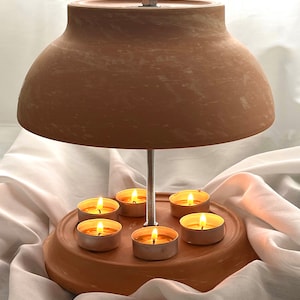 Puede incluir: Un calentador de velas de terracota con una parte superior en forma de cúpula y una base circular. Seis velas de té se colocan en la base, creando un brillo cálido.