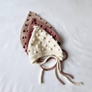 Puede incluir: Tres gorros de bebé tejidos en crema, malva y topo, cada uno adornado con bolitas texturizadas. Los gorros tienen lazos para asegurar debajo de la barbilla. Los sombreros están dispuestos en una composición en capas, mostrando los diferentes colores y texturas.
