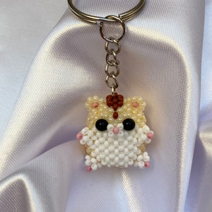 May include: A white beaded hamster keyring with pink accents and black eyes. The hamster is attached to a silver key ring.