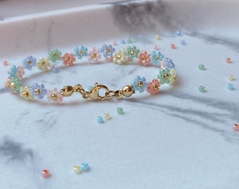 May include: A delicate bracelet made with pastel-colored flower beads and gold-toned clasps. The bracelet is on a white marble surface.
