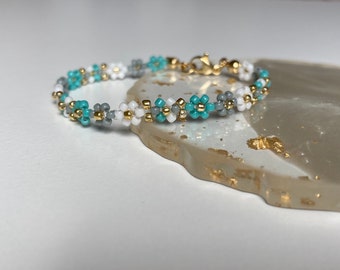 May include: A delicate beaded bracelet with a gold clasp. The bracelet features a repeating pattern of turquoise, white, and grey beads.