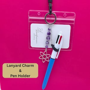 May include: A lanyard charm and pen holder set against a pink background. The charm features purple beads and a silver starburst pendant. A multi-color pen is held by a black rubber grip. The text "Lanyard Charm & Pen Holder" is in a tan box.